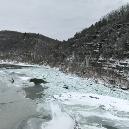 A snowy landscape with a frozen river and icy patches, surrounded by wooded hills under a cloudy sky.