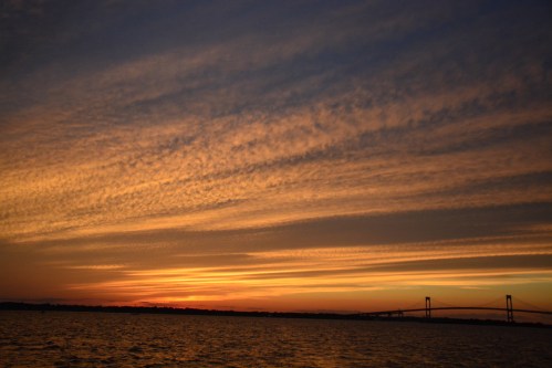 A vibrant sunset over water, with colorful clouds and a bridge silhouetted against the horizon.
