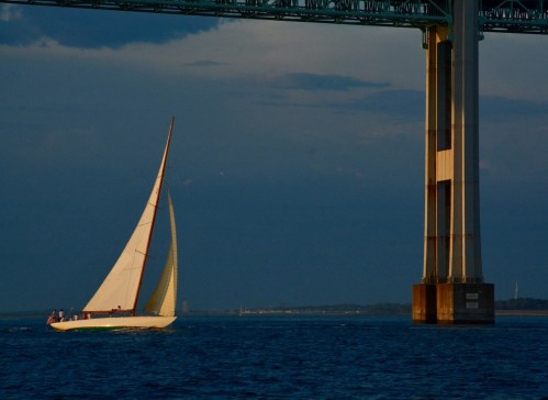 A sailboat glides under a bridge, with calm waters and a dramatic sky in the background.