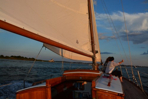 A sailboat glides through water at sunset, with two people enjoying the view from the deck.