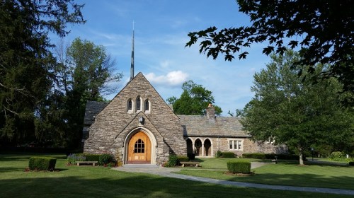 A stone church with a tall spire, surrounded by green trees and a well-maintained lawn under a blue sky.