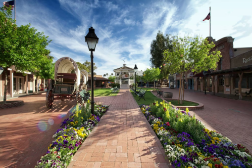 A scenic pathway lined with flowers, leading to a historic building and vintage wagons under a blue sky.
