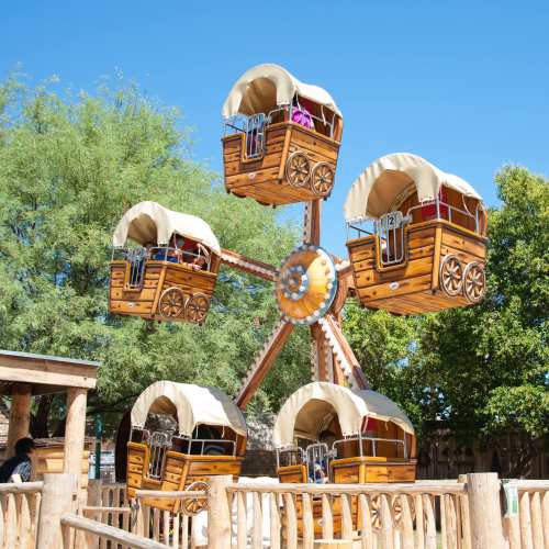 A whimsical Ferris wheel with wagon-style seats against a clear blue sky and green trees.