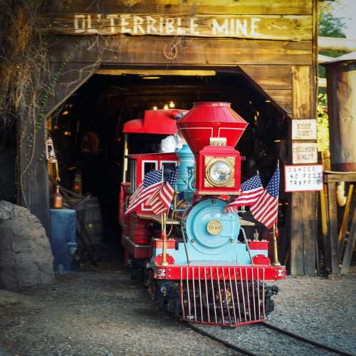 A colorful steam train with American flags exits a rustic mine entrance labeled "Ol' Terrible Mine."