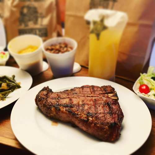 A grilled steak on a white plate, surrounded by sides of salad, beans, greens, and a drink in the background.