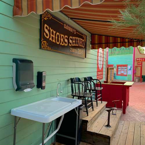 A colorful outdoor shoe-shining station with a sign, sink, and chairs, set against a vibrant backdrop.