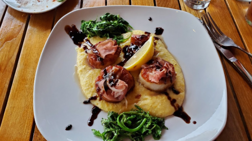 A plate of scallops topped with prosciutto, served on creamy polenta with a lemon wedge and greens.