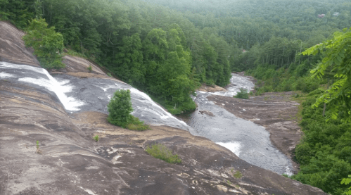 A scenic view of a waterfall cascading down smooth rocks into a winding river, surrounded by lush green trees.