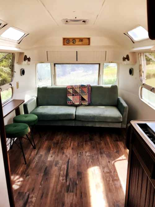 Cozy interior of a vintage camper with a green sofa, colorful quilt, and wooden accents, surrounded by windows.
