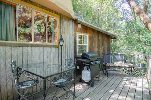 A wooden deck with a glass table, chairs, and a grill, surrounded by trees and greenery.