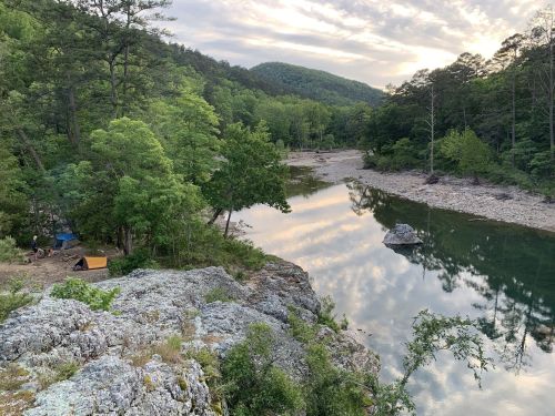A serene river scene surrounded by lush trees and mountains, with a campsite visible on the riverbank.