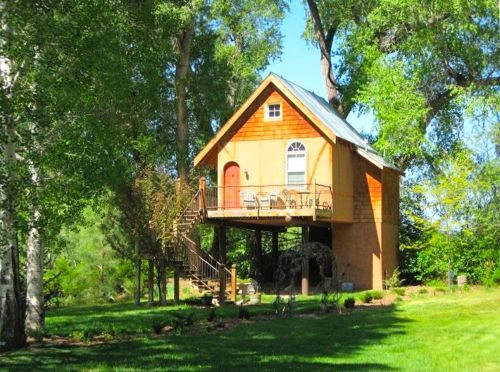 A charming orange treehouse with a porch, surrounded by lush greenery and tall trees.