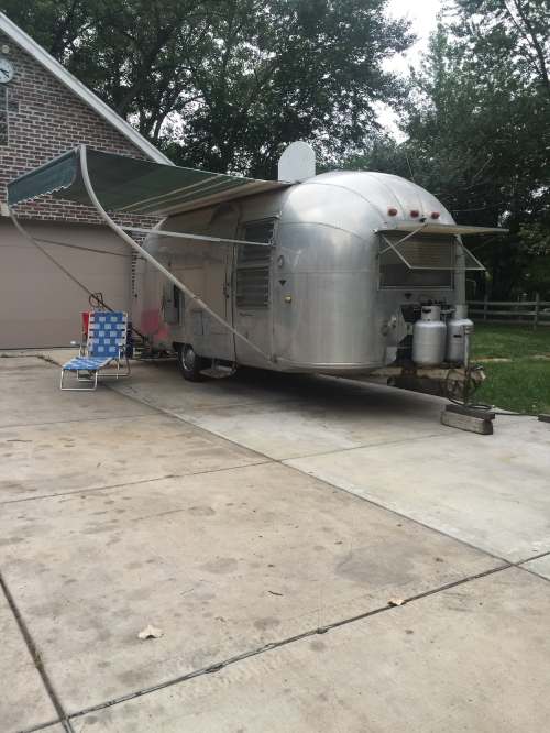 A vintage Airstream trailer parked beside a house, with an awning extended and a folding chair nearby.