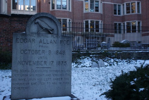 Gravestone of Edgar Allan Poe, detailing his burial dates, surrounded by snow and a historic building in the background.
