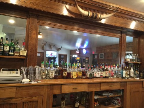 A wooden bar with various bottles of liquor displayed, featuring a rustic decor and mounted deer heads in the background.