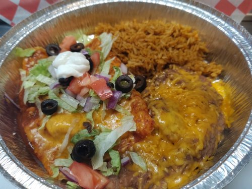 A foil bowl filled with rice, enchiladas topped with cheese, lettuce, tomatoes, olives, and sour cream.