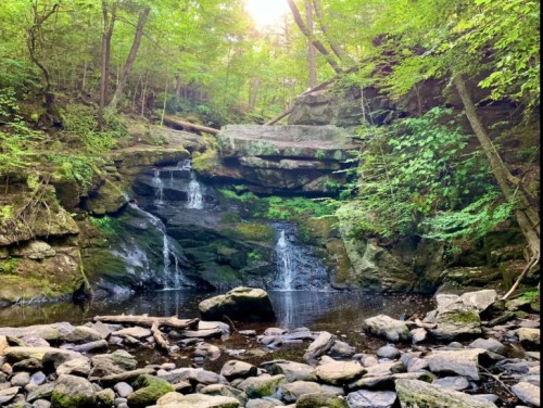 A serene waterfall cascades over moss-covered rocks, surrounded by lush green trees and a rocky riverbed.
