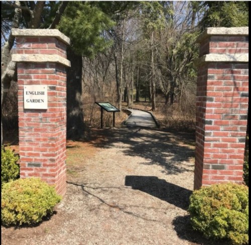 Entrance to an English garden, featuring brick pillars and a wooden walkway surrounded by trees and greenery.