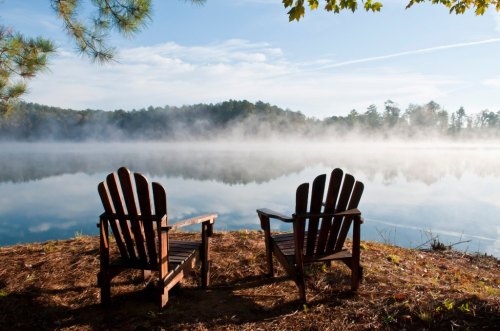 Two wooden Adirondack chairs facing a misty lake, surrounded by trees under a clear blue sky.