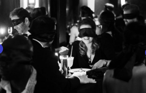 A group of people at a dimly lit restaurant, all wearing blindfolds, enjoying a unique dining experience.