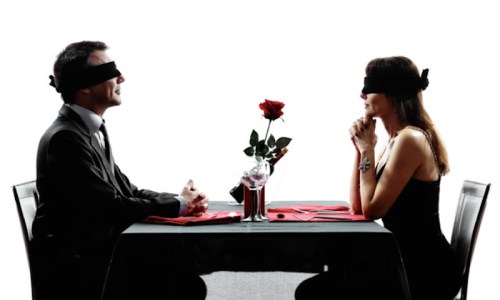 A man and woman sit at a table, both blindfolded, with a rose between them on a black tablecloth.