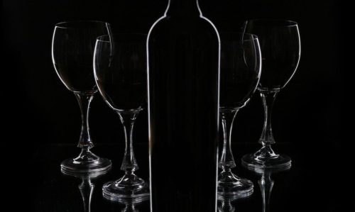 A silhouette of a wine bottle in front of five empty wine glasses against a black background.