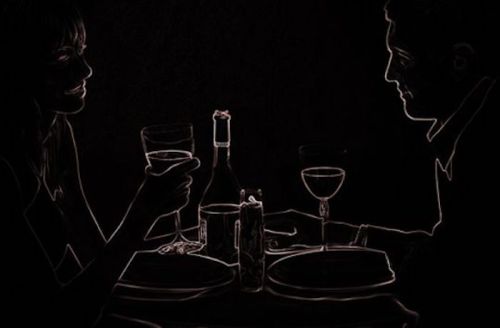 A couple enjoys a candlelit dinner, holding glasses and smiling at each other in a dimly lit setting.