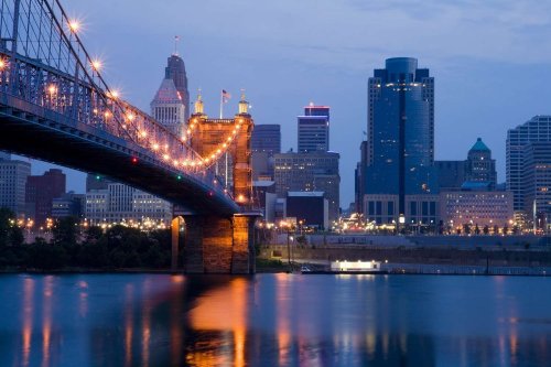 A city skyline at dusk, featuring a bridge illuminated with lights over a calm river.