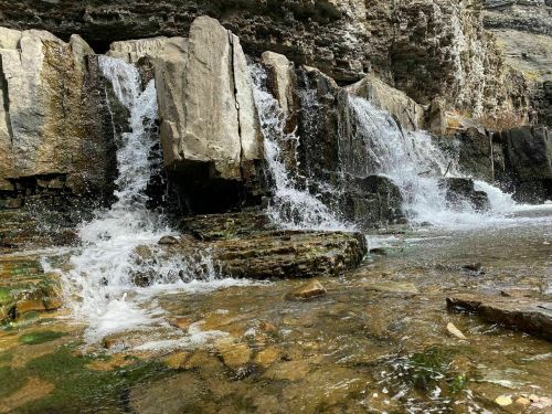 A small waterfall cascades over rocky ledges into a clear stream, surrounded by rugged cliffs and natural greenery.