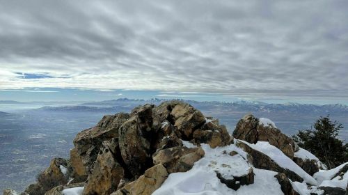 A rocky mountain peak covered in snow, with a cloudy sky and distant mountains visible in the background.