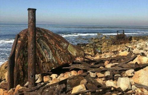 Rusty metal remnants and weathered rocks along a shoreline, with calm waves and a clear blue sky in the background.