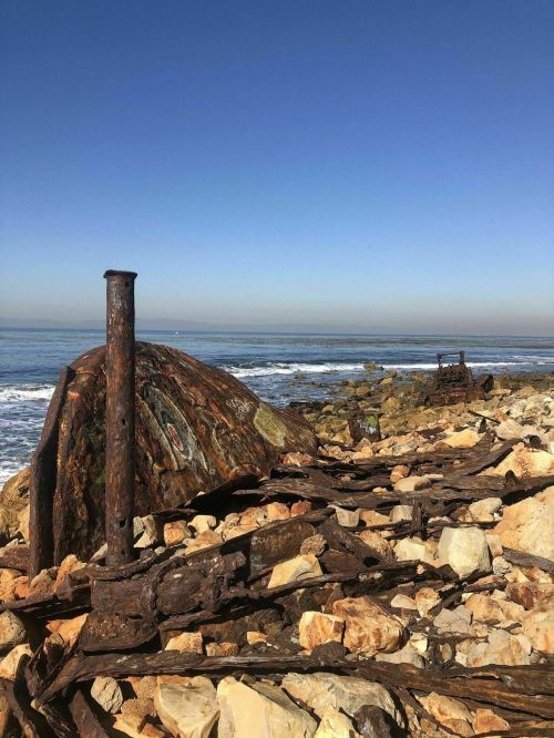 Rusty remnants of a shipwreck on rocky shore, with calm waves and clear blue sky in the background.