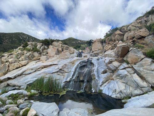 A serene waterfall cascades over rocky terrain, surrounded by lush greenery and a cloudy blue sky.