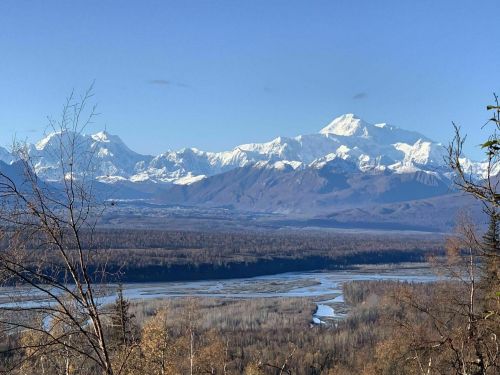 Snow-capped mountains rise above a valley with a winding river, under a clear blue sky. Trees frame the foreground.