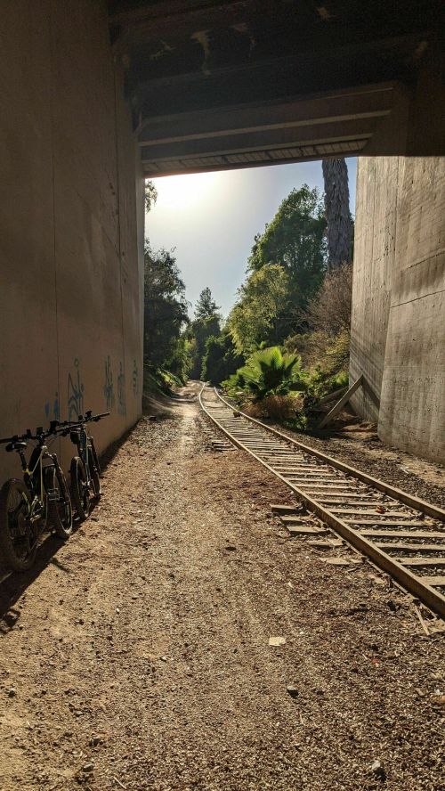 A view of a dirt path and railway tracks under a bridge, with bicycles parked on the side and greenery in the background.