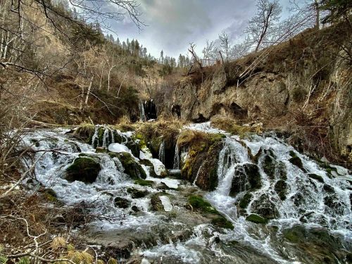 A serene waterfall cascades over rocks, surrounded by bare trees and rugged terrain under a cloudy sky.