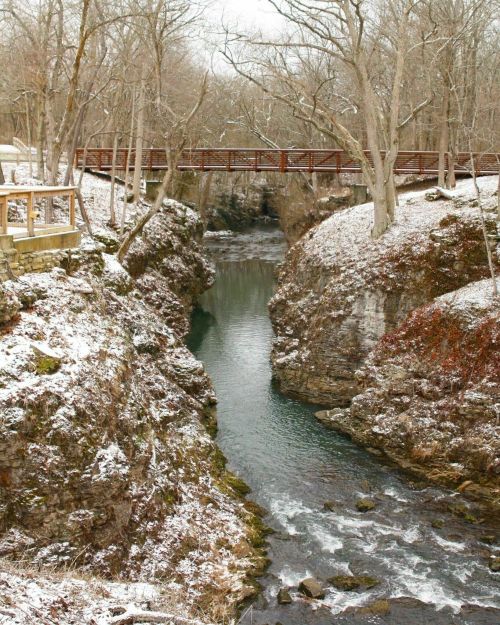 A snowy landscape featuring a narrow river flowing through rocky cliffs, with a wooden bridge overhead.