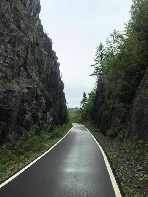 A winding road between steep, rocky cliffs, surrounded by lush greenery under a cloudy sky.