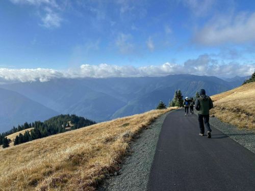 Hikers walking on a path through a mountainous landscape with clouds and blue skies in the background.