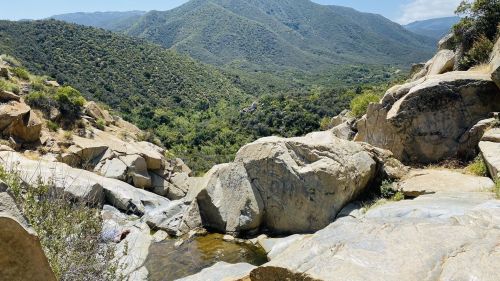 A rocky landscape with a small pool of water, surrounded by lush green hills and mountains under a clear blue sky.