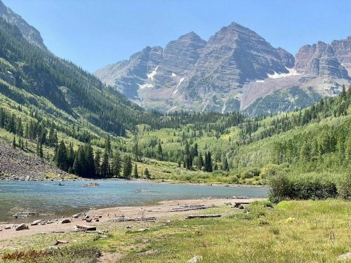 A serene landscape featuring a clear lake surrounded by lush greenery and towering mountains under a blue sky.
