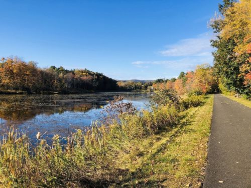 A serene river scene with colorful autumn foliage and a paved path alongside the water under a clear blue sky.