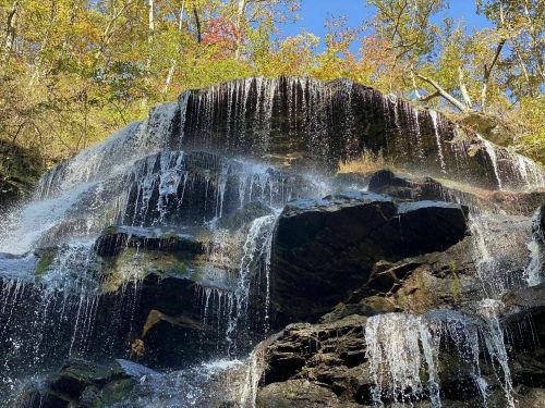 A cascading waterfall flows over rocky cliffs, surrounded by vibrant autumn foliage and a clear blue sky.