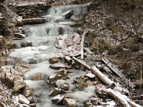 A frozen stream flows over rocks, surrounded by snow-covered ground and bare trees in a winter landscape.