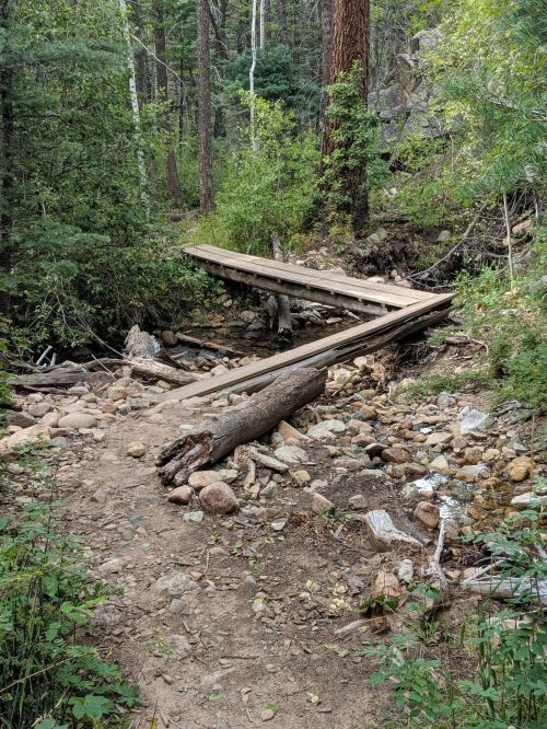A wooden bridge crosses a rocky stream in a forested area, surrounded by trees and greenery.