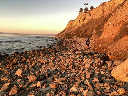 A person walks along a rocky beach at sunset, with cliffs and palm trees in the background.