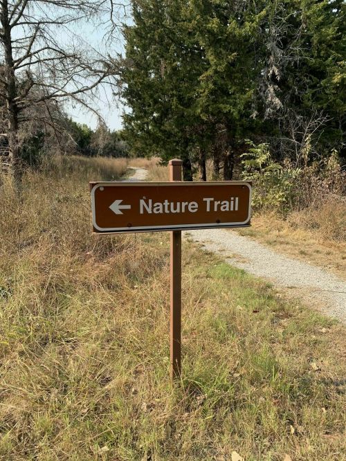 A brown sign pointing left, indicating the direction of a nature trail, surrounded by trees and tall grass.