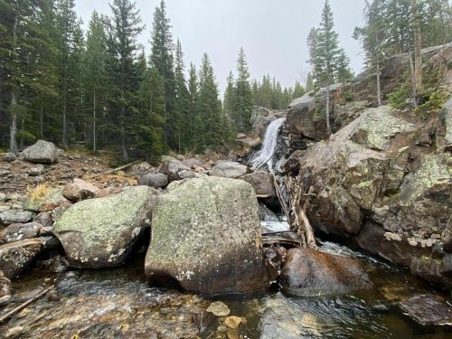 A serene waterfall cascades over rocks, surrounded by lush green trees in a misty forest setting.