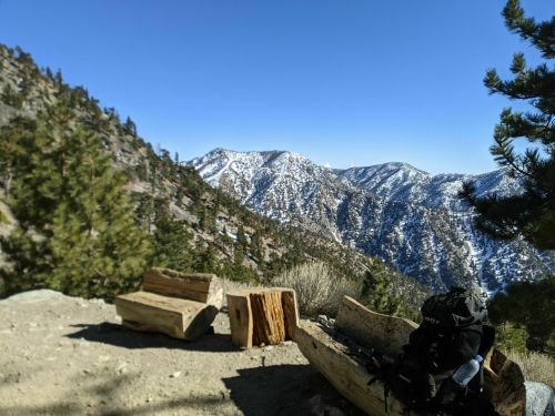Scenic mountain view with snow-capped peaks, wooden benches, and a backpack in a sunny outdoor setting.