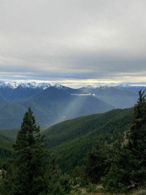 A panoramic view of mountains with snow-capped peaks, lush green valleys, and a cloudy sky. Sunlight breaks through the clouds.